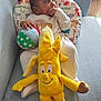 baby, child, plush_toy, yellow_banana, ball, couch, pillow, animal_print, striped_clothing, hand, face, sitting, indoors, soft_toy, infant, looking_away, furniture, cute, comfortable, young_child
