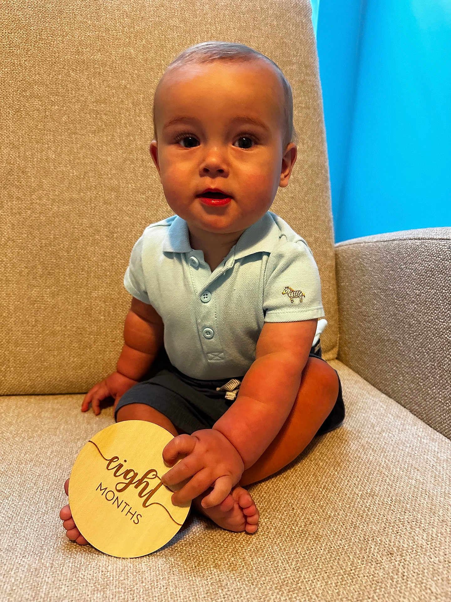 Chayce is registered to the contest to win money with this photo: baby, infant, child, sitting, polo_shirt, shorts, couch, beige, wooden_sign, eight_months, cute, portrait, indoors, soft_lighting, curious, hand, feet, baby_toys, smiling, young_child