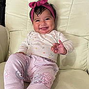 Aurora is registered to the contest to win money with this photo: baby, smiling, headband, pink, pajamas, patterned_clothing, cushion, chair, indoors, comfort, happy, person, infant, cute, child, sitting, hand_gesture, face, hair, relaxed