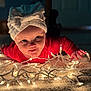baby, child, face, headwear, bow, lights, string_lights, carpet, indoors, red_clothing, smiling, curious, glowing, portrait, crawling, closeup, soft_light, holiday, festive, warm