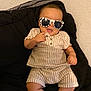 baby, child, sunglasses, striped_clothing, cushion, black_background, indoor, smiling, cute, fashion, infant, relaxed, person, portrait, shorts, shirt, comfort, seated, hand, playful