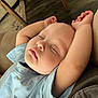 baby, sleeping, arms_up, blue_shirt, infant, peaceful, face, closed_eyes, chubby_cheeks, indoor, couch, wooden_floor, relaxing, soft_lighting, portrait, child, toddler, casual_clothing, home, resting