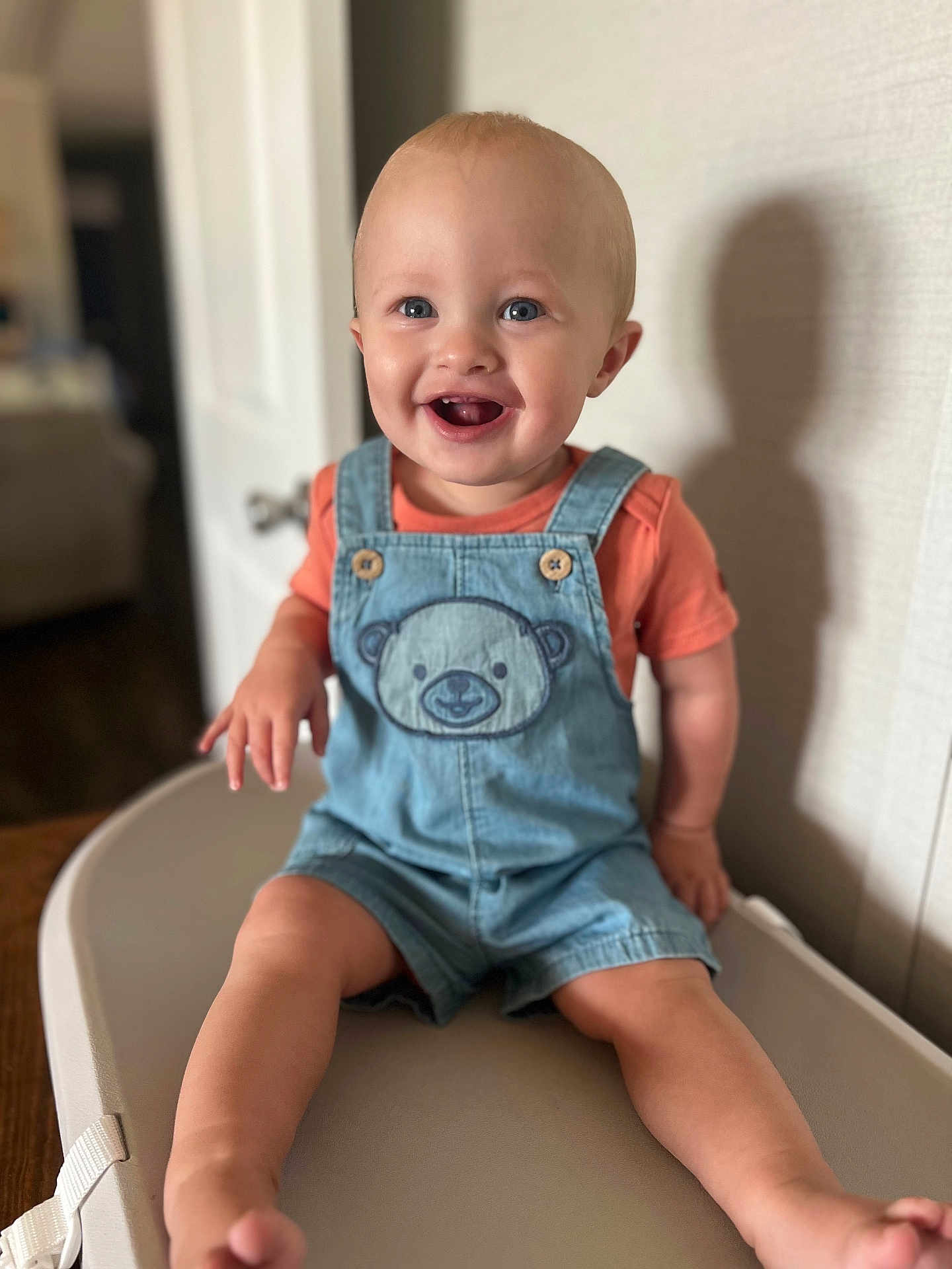 Sonny is registered to the contest to win money with this photo: baby, child, smiling, denim_overalls, teddy_bear_patch, orange_shirt, high_chair, indoor, happy, cute, portrait, blond_hair, blue_eyes, shadow, furniture, person, seated, skin, face, table