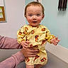 baby, barefoot, child, cute, hands, human, indoor, infant, medical_room, person, rosy_cheeks, short_hair, skin, smiling, standing, support, table, tiger_print, yellow_gown, young