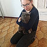 affection, animal, black_clothing, casual, cat, comfort, door, floor, glasses, gray_cat, home, hugging, indoor, kneeling, long_hair, person, pet, tile_floor, watch, young_person