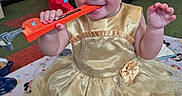 Delylah is registered to the contest to win money with this photo: baby, child, dress, golden_dress, toy, orange_toy, fire_truck_toy, playroom, carpet, blanket, sitting, smiling, happy, indoor, cute, infant, person, earrings, foot, hand