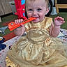 baby, child, dress, golden_dress, toy, orange_toy, fire_truck_toy, playroom, carpet, blanket, sitting, smiling, happy, indoor, cute, infant, person, earrings, foot, hand
