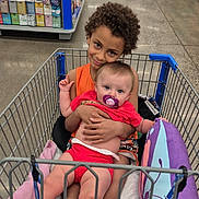 Delylah joined the competition — help win amazing prizes! child, baby, shopping_cart, store, aisle, floor, pillow, pacifier, clothing, person, retail, smiling, hugging, curly_hair, shopping, sitting, colorful, indoor, happy, cute