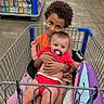 child, baby, shopping_cart, store, aisle, floor, pillow, pacifier, clothing, person, retail, smiling, hugging, curly_hair, shopping, sitting, colorful, indoor, happy, cute