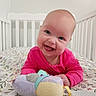 baby, smiling, crib, toy, plush_toy, pink_clothing, floral_pattern, infant, happy, child, nursery, bed, head, face, person, cute, indoor, playful, toddler, lying_down