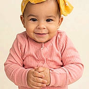 Skye is registered to the contest to win money with this photo: toddler, child, baby, smile, face, headband, yellow_bow, pink_clothing, long_sleeve, hands, portrait, cute, happy, infant, studio, neutral_background, young_child, person, indoors, fashion