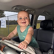 Johnny is registered to the contest to win money with this photo: baby, child, smiling, car_seat, vehicle_interior, blue_eyes, green_tank_top, happy, infant, seat, hand, window, daylight, fence, outdoor_view, portrait, cute, young_child, car, leisure