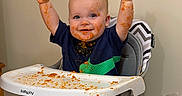 Johnny joined the competition — help win amazing prizes! baby, high_chair, messy, food, orange_food, happy, smiling, child, mealtime, indoor, plate, bowl, table, clothing, face, arms_up, cute, person, feeding, home