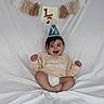 Al is registered to the contest to win money with this photo: baby, celebration, child, clothing, cute, decorations, fabric, half_birthday, happy, indoor, infant, milestone, party_hat, person, portrait, seated, smiling, sweater, tassels, white_background