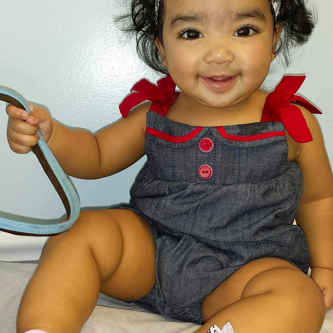 Zuneira is registered to the contest to win money with this photo: baby, child, cute, denim_dress, footwear, hand, happy, headband, indoor, infant, person, playful, portrait, red_flower, sitting, skin, smiling, socks, white_background, young_child