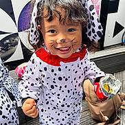 Skylar joined the competition — help win amazing prizes! toddler, child, costume, dalmatian, face_paint, smile, snack, chips, outdoor, wooden_deck, black_and_white, pattern, floppy_ears, cute, happy, clothing, portrait, person, fun, playful
