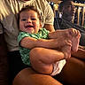 baby, child, infant, smiling, happy, portrait, family, adult, parent, sitting, green_outfit, diaper, bare_feet, arms, legs, indoor, evening, string_lights, warm_lighting, candid