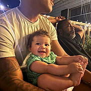 Kiyan Alan is registered to the contest to win money with this photo: baby, child, infant, smiling, outdoor, night, patio, adults, family, portrait, sitting, green_outfit, bare_feet, warm_lighting, string_lights, window, tattoo, beard, casual, happy