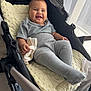 baby, stroller, smiling, gray_clothing, ribbed_leggings, blanket, indoor, natural_light, curtains, child, happy, infant, hand, fabric, face, cheeks, clothing, soft_toy, candid, relaxing