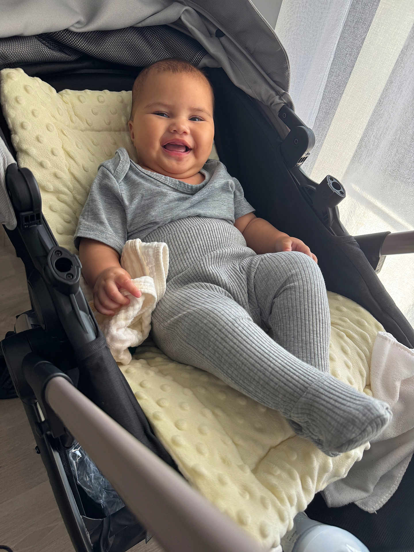 Arofahere participe au concours pour gagner de l'argent avec cette photo : baby, stroller, smiling, gray_clothing, ribbed_leggings, blanket, indoor, natural_light, curtains, child, happy, infant, hand, fabric, face, cheeks, clothing, soft_toy, candid, relaxing