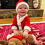 baby, blanket, child, christmas, cute, festive, fireplace, floor, happy, hat, holiday_decor, indoor, person, plush_toy, red_clothing, reindeer_toy, santa_claus_costume, seasonal, smiling, toy