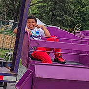 Jayvion is registered to the contest to win money with this photo: child, smiling, amusement_park, ride, purple, red_pants, sneakers, outdoor, trees, fence, playground,  amusement_ride, happy, portrait, seated, camera, family_fun, metal_platform, daytime, recreation