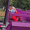 child, smiling, amusement_park, ride, purple, red_pants, sneakers, outdoor, trees, fence, playground,  amusement_ride, happy, portrait, seated, camera, family_fun, metal_platform, daytime, recreation