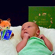 Oliver is registered to the contest to win money with this photo: baby, infant, child, portrait, lying_down, green_clothing, blanket, plush_toy, fox_toy, robot_toy, uno_cards, playing_cards, book, nursery, cozy, indoor, awake, hand_to_mouth, colorful, smiling_toy