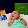 awake, baby, blanket, book, child, colorful, cozy, fox_toy, green_clothing, hand_to_mouth, indoor, infant, lying_down, nursery, playing_cards, plush_toy, portrait, robot_toy, smiling_toy, uno_cards
