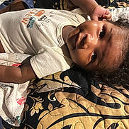 Legend is registered to the contest to win money with this photo: toddler, child, baby, face, expression, curly_hair, white_shirt, colorful_print, blanket, patterned_blanket, indoor, lying_down, portrait, skin, hand, cute, young_child, soft_light, close_up, infant