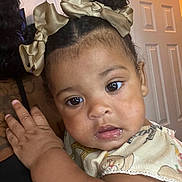 London is registered to the contest to win money with this photo: baby, child, face, curly_hair, hair_bow, indoor, person, closeup, expression, skin, hand, clothing, shoulder, portrait, cute, young_child, looking, curious, light, warm