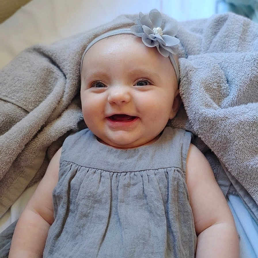 Journey is registered to the contest to win money with this photo: baby, smiling, headband, flower, blanket, dress, child, infant, happy, portrait, cute, indoors, cozy, skin, face, person, apparel, soft, lying_down, toddler