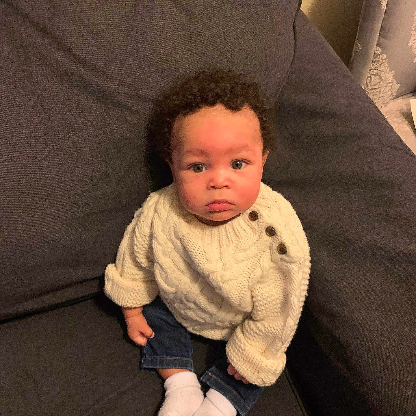 Tyshawn joined the competition — help win amazing prizes! baby, child, curly_hair, sweater, white_socks, jeans, couch, indoor, portrait, seated, looking_up, cute, cozy, knitwear, furniture, person, young_child, home, casual_clothing, expression