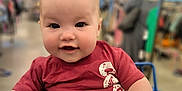 Adalynn joined the competition — help win amazing prizes! baby, child, shopping_cart, toy, red_shirt, smiling, indoor, store, aisle, blurred_background, person, cute, happy, infant, face, hand, clothing, retail, seatbelt, shopping