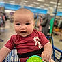 baby, child, shopping_cart, toy, red_shirt, smiling, indoor, store, aisle, blurred_background, person, cute, happy, infant, face, hand, clothing, retail, seatbelt, shopping