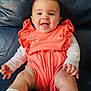 baby, infant, smiling, sitting, coral_outfit, ruffles, white_socks, lace, chubby_legs, happy, cute, leather_couch, indoor, child, portrait, young, person, hands, face, bright_clothing