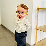 Sacha a rejoint le concours — aidez-le/la à gagner de superbes lots ! child, glasses, white_shirt, dark_pants, sneakers, indoor, tile_floor, wooden_shelf, playful, happy, standing, young_boy, casual_clothing, short_hair, expression, brown_hair, smiling, person, home_interior, light_wall