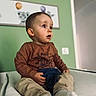 toddler, child, indoor, sitting, brown_shirt, beige_pants, curious, soft_surface, green_wall, framed_picture, animal_print, cute, young_child, portrait, casual_clothing, white_socks, home_interior, expression, person, baby