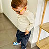 toddler, child, glasses, sneakers, standing, indoor, floor, tile_floor, shelf, white_wall, clothing, long_sleeve, pants, casual_wear, looking_down, hair, person, minimalist, home, footwear