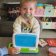 Dario a rejoint le concours — aidez-le/la à gagner de superbes lots ! toddler, child, pacifier, toy_laptop, table, books, toys, smile, indoor, living_room, couch, storage_boxes, colorful, happy, play, person, furniture, young_child, clothing, hand