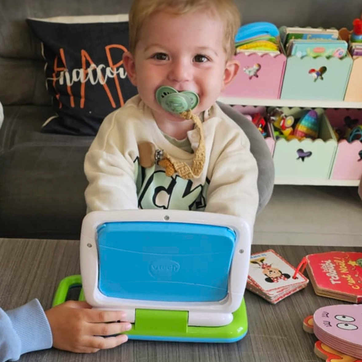 Dario a rejoint le concours — aidez-le/la à gagner de superbes lots ! books, child, clothing, colorful, couch, furniture, hand, happy, indoor, living_room, pacifier, person, play, smile, storage_boxes, table, toddler, toy_laptop, toys, young_child