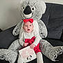 baby, infant, stuffed_animal, koala, plush_toy, bow, red_bow, milestone_card, four_months, surprised_expression, wide_eyes, sofa, couch, sitting, white_outfit, red_skirt, tights, plush_legs, portrait, cozy