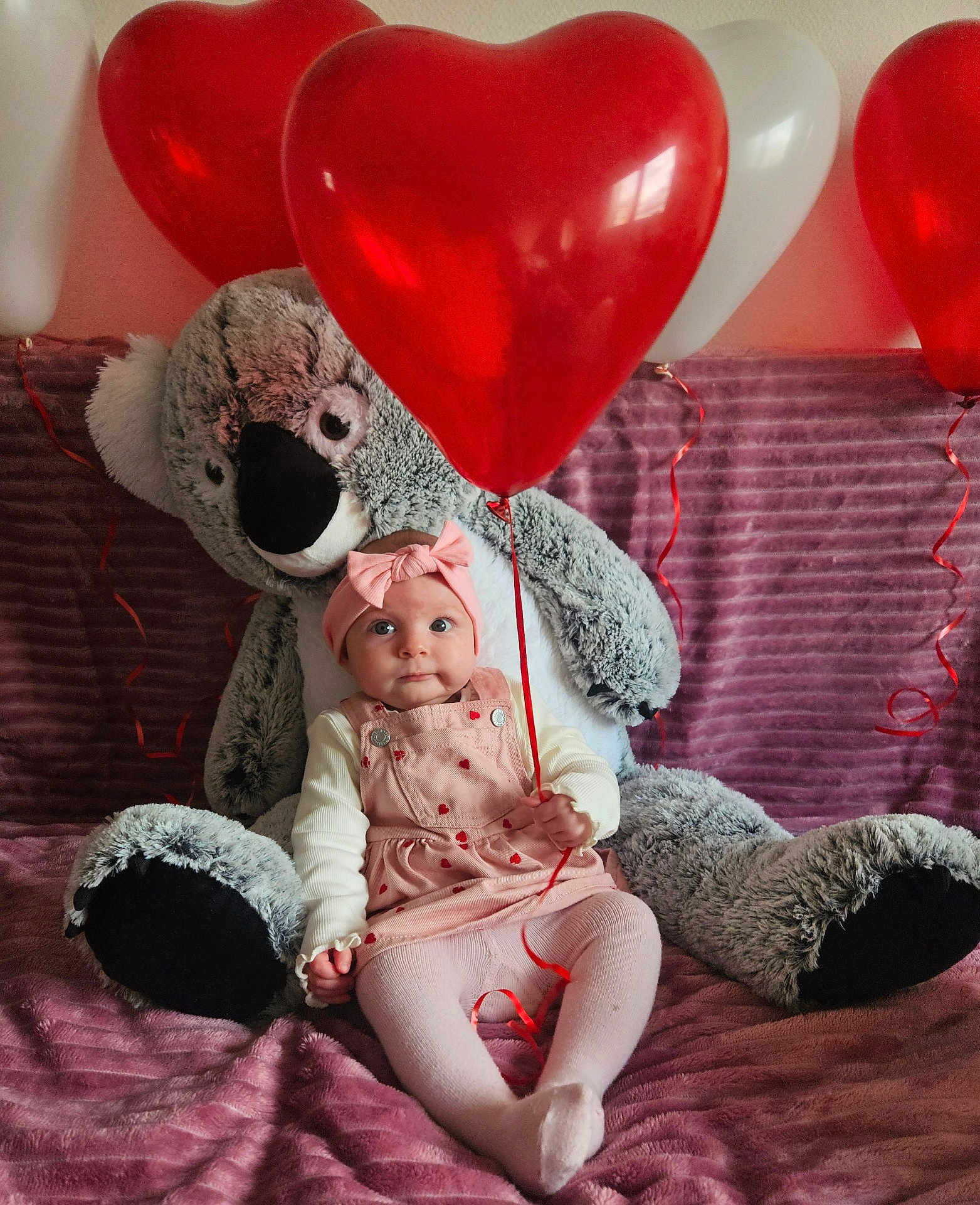 Ambre a rejoint le concours — aidez-le/la à gagner de superbes lots ! baby, infant, child, stuffed_koala, stuffed_animal, plush_toy, heart_balloon, balloons, headband, bow, pink_dress, tights, sitting, blanket, bed, large_toy, adorable, portrait, indoor, ribbon