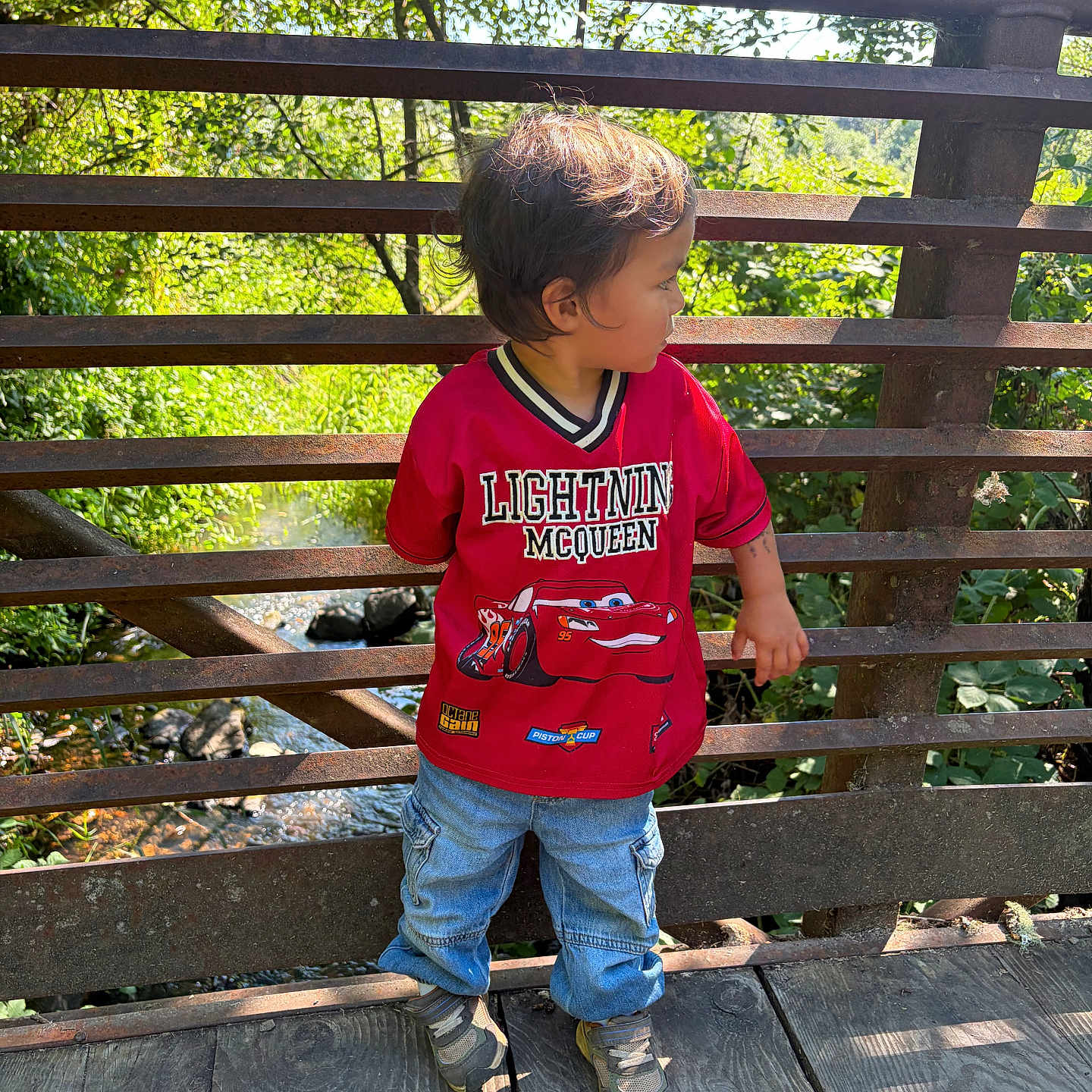 Marcelo is registered to the contest to win money with this photo: toddler, child, red_shirt, lightning_mcqueen, jeans, sneakers, wooden_bridge, metal_railings, outdoor, greenery, trees, creek, daylight, nature, curious, standing, casual_clothing, summer, sunlight, portrait