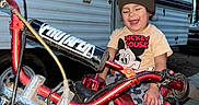 Marcelo is registered to the contest to win money with this photo: child, toddler, dirt_bike, motorcycle, beanie, mickey_mouse, tshirt, smiling, outdoor, camper, vehicle, helmet_absent, dirt, wheel, handlebar, exhaust, person, happy, clothing, play