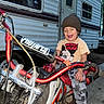 child, toddler, dirt_bike, motorcycle, beanie, mickey_mouse, tshirt, smiling, outdoor, camper, vehicle, helmet_absent, dirt, wheel, handlebar, exhaust, person, happy, clothing, play
