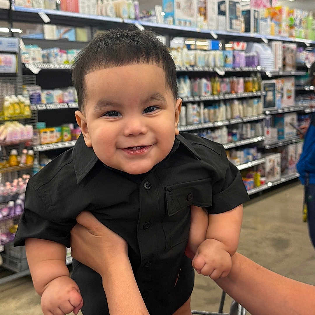 Emiliano joined the competition — help win amazing prizes! baby, child, infant, smiling, happy, portrait, black_outfit, short_hair, supermarket, grocery_store, shopping_cart, store_aisle, shelves, retail, indoors, shopping, adult_hands, standing, closeup, family