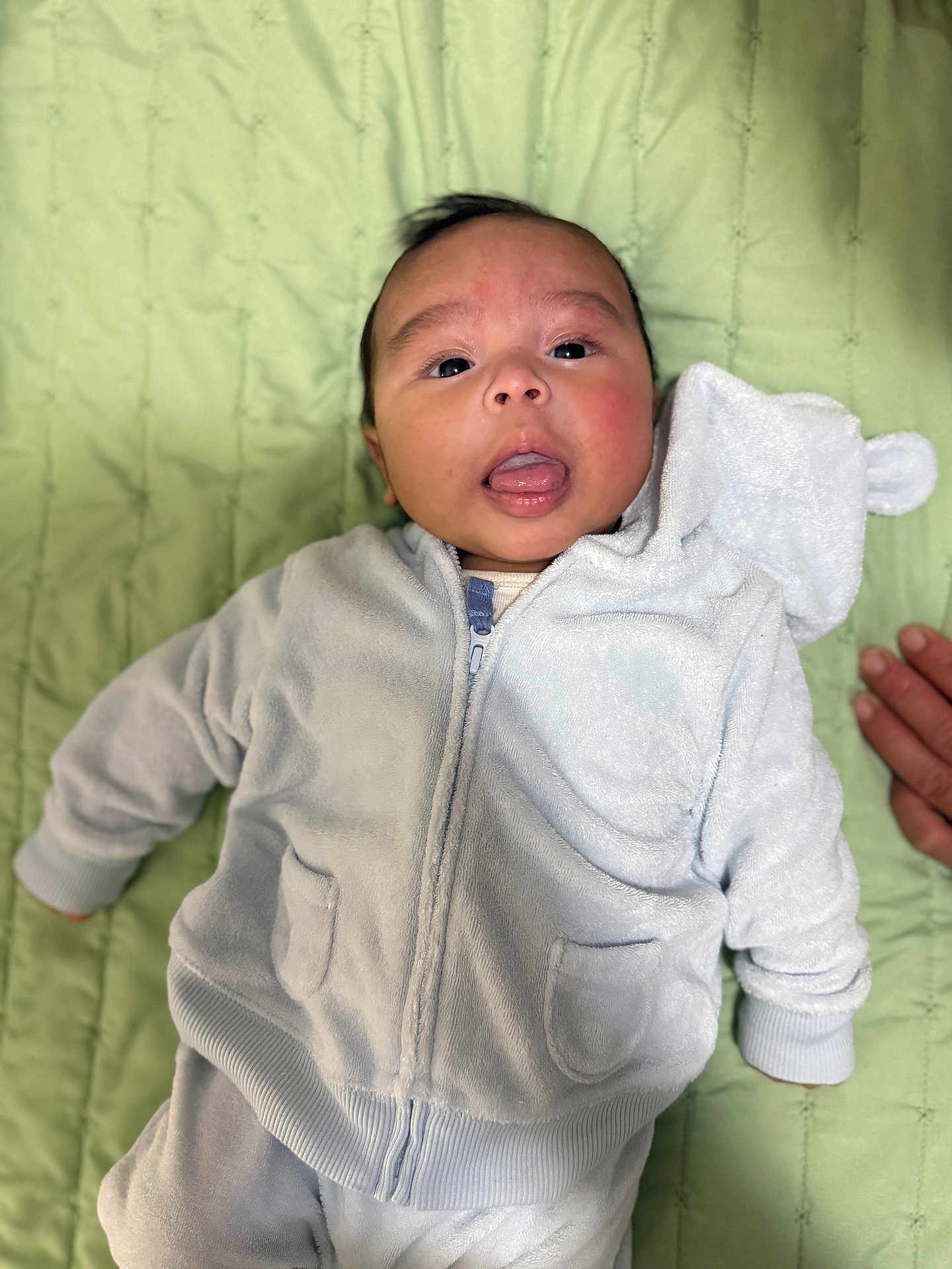 Mariano joined the competition — help win amazing prizes! baby, infant, child, hoodie, white_clothing, blanket, green_blanket, face, tongue_out, hand, cute, soft_texture, lying_down, portrait, young_child, apparel, zipped_jacket, pocket, indoor, person