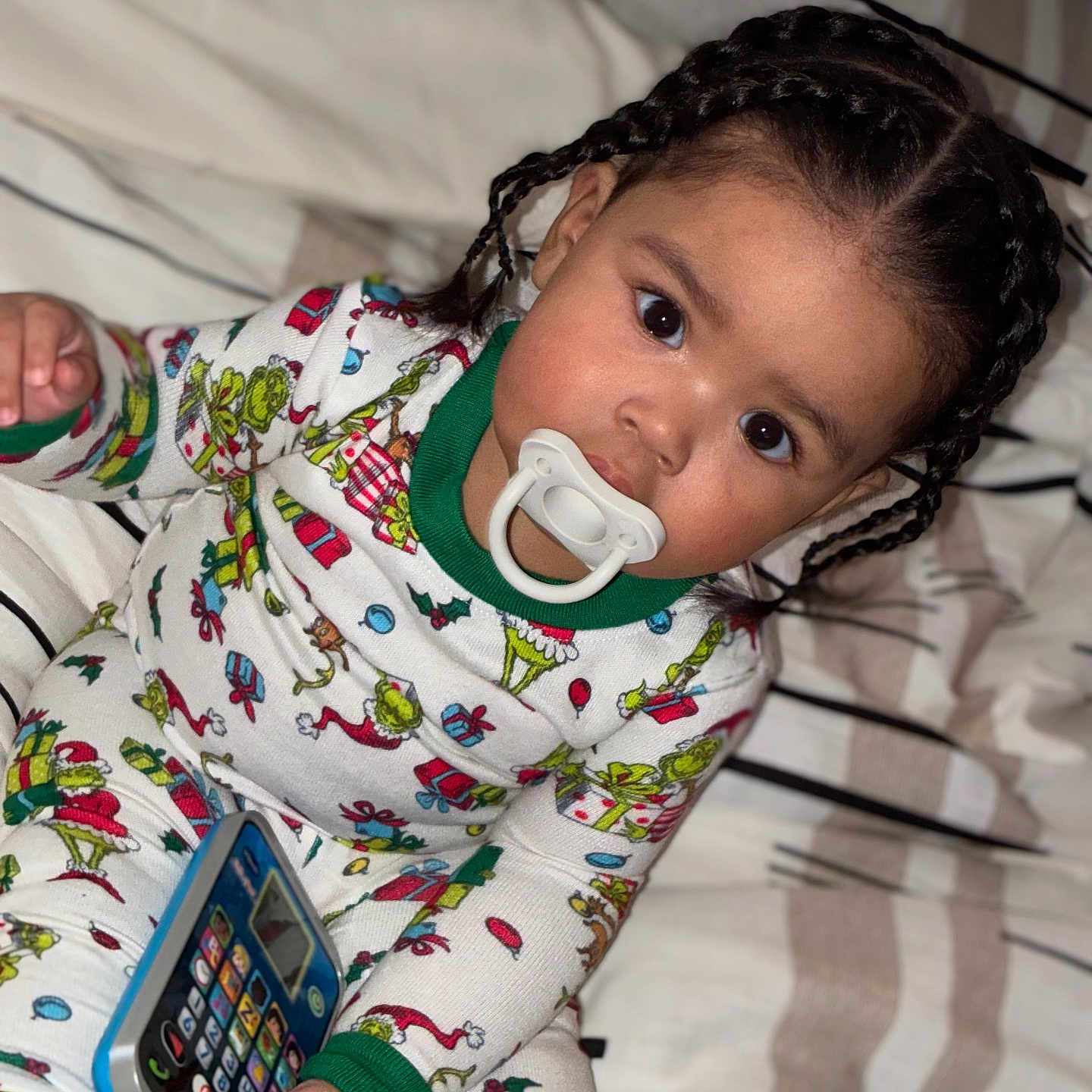 Kartier is registered to the contest to win money with this photo: bed, blanket, braided_hair, child, colorful, comfort, cute, holding, indoor, pacifier, pajamas, person, play, portrait, sleepwear, smiling, striped, toddler, toy_phone, young_child