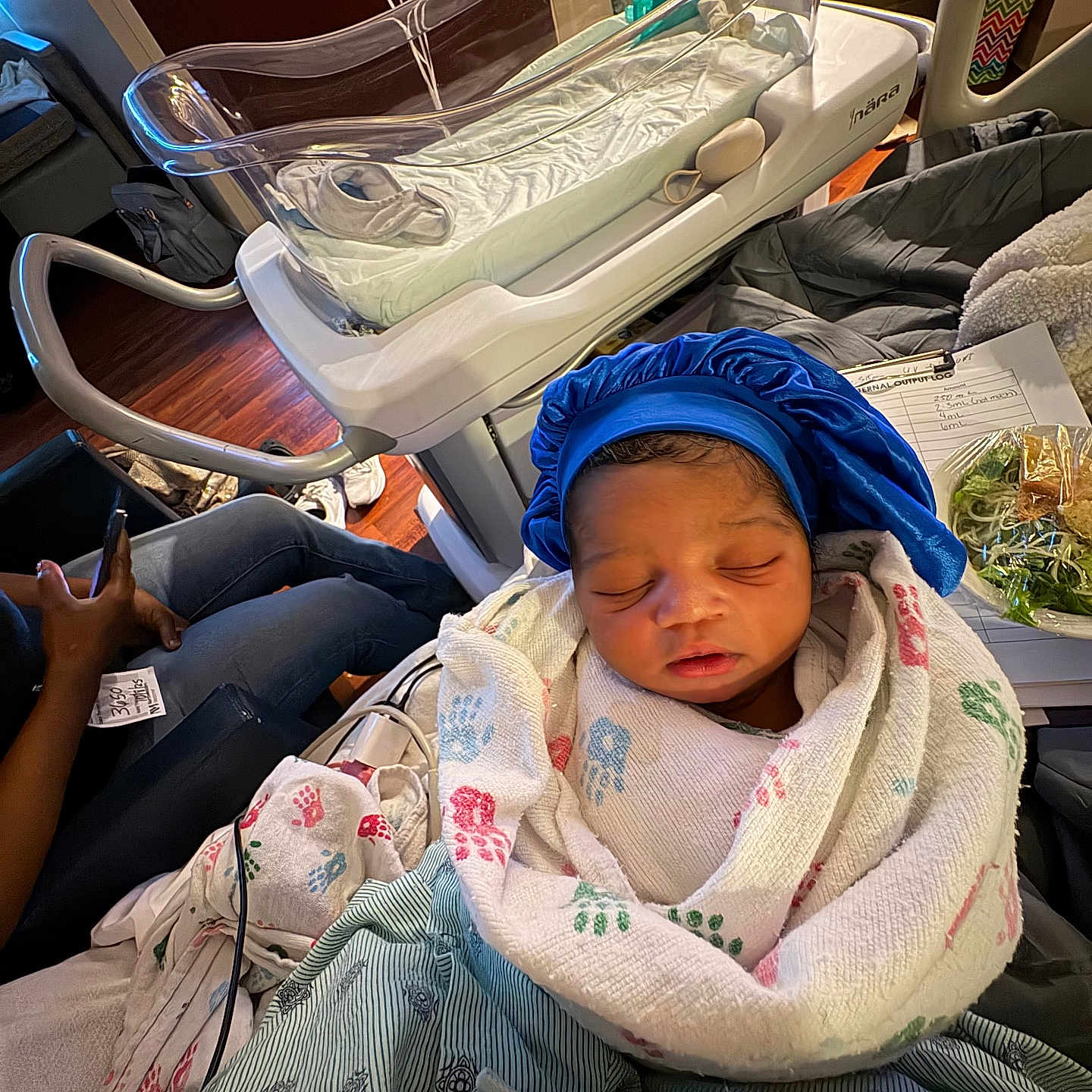 Nyaire is registered to the contest to win money with this photo: newborn, baby, hospital, blanket, bassinet, bed, person, blue_bonnet, sleeping, infant, medical, resting, indoor, clothing, furniture, wooden_floor, hand, phone, paper, peaceful